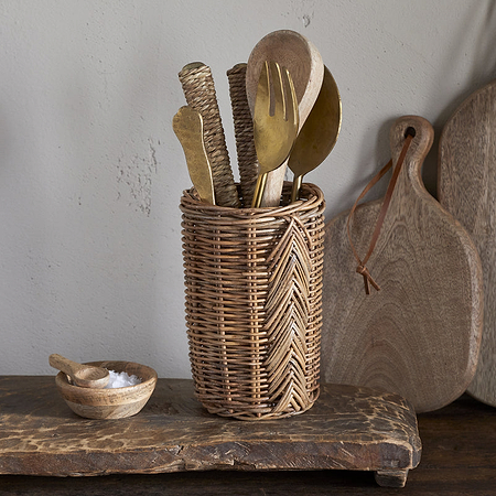 The nkuku Bersama Rattan Utensil Holder - Natural, filled with assorted wooden and brass cutlery, rests on a rustic surface next to a small wooden salt bowl. Wooden cutting boards hang on the wall in the background.