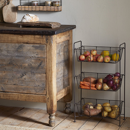 A rustic wooden cabinet sits on a wood floor beside the nkuku Inkollu Three Tier Storage Stand in antiqued brass, filled with apples, carrots, onions, and potatoes. A loaf of bread is on a cutting board atop the cabinet; kitchen items and greenery are visible.
