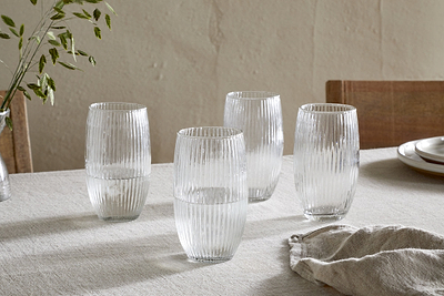 A set of four nkuku Otuba Highball Clear glasses is arranged on a light beige tablecloth, each half-filled with water. Wooden chairs, stacked plates, a cloth napkin, and a small vase of greenery complete the elegant table setting.