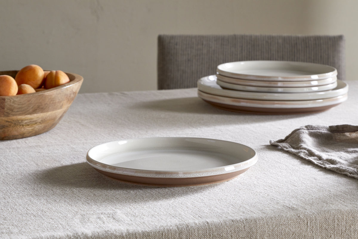 A stack of ceramic plates and the nkuku Edo Side Plate - Terracotta rest on a textured beige tablecloth. To the left, a wooden bowl holds apricots, while a gray napkin completes this elegant minimalist tableware arrangement.