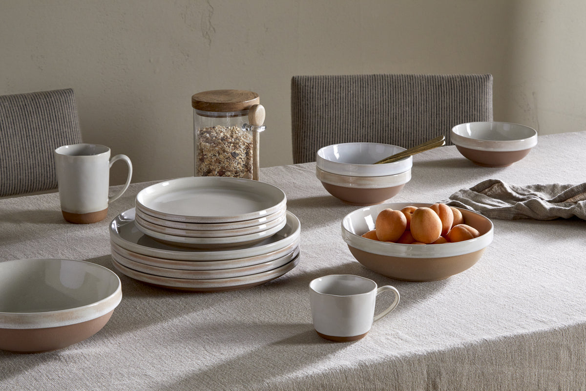 A table set with minimalist beige ceramic plates, bowls, and cups—some stacked. A large nkuku Edo Cereal Bowl in Terracotta holds apricots. There’s a glass jar of oats, striped napkin, and neutral chairs on a light tablecloth.