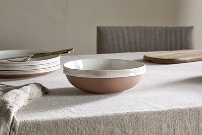 A handcrafted nkuku Edo Pasta Bowl - Terracotta with a white interior and terracotta exterior rests on a light linen tablecloth, surrounded by minimalist tableware—stacked plates, fork, wooden board—and a gray upholstered chair in a simple setting.
