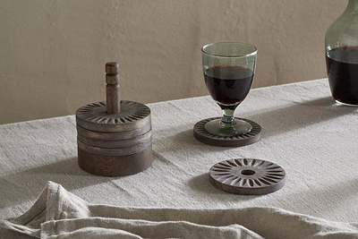 A set of nkuku Eisha Mango Wood Coasters (dark brown, set of 6) with hand-carved edges sits on a beige tablecloth; one holds a glass of red wine, while a matching carafe nearby enhances the rustic charm under soft lighting.