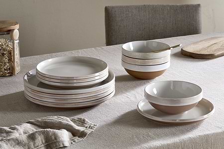 A neutral-toned table features the nkuku Edo Dinnerware Set in terracotta—stacked ceramic plates, bowls, and cups. A wooden cutting board, jar of grains, and folded napkin complete the rustic arrangement.