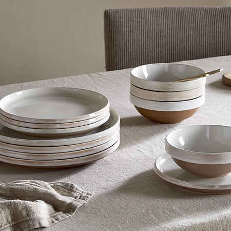 A neutral-toned table features the nkuku Edo Dinnerware Set in terracotta—stacked ceramic plates, bowls, and cups. A wooden cutting board, jar of grains, and folded napkin complete the rustic arrangement.