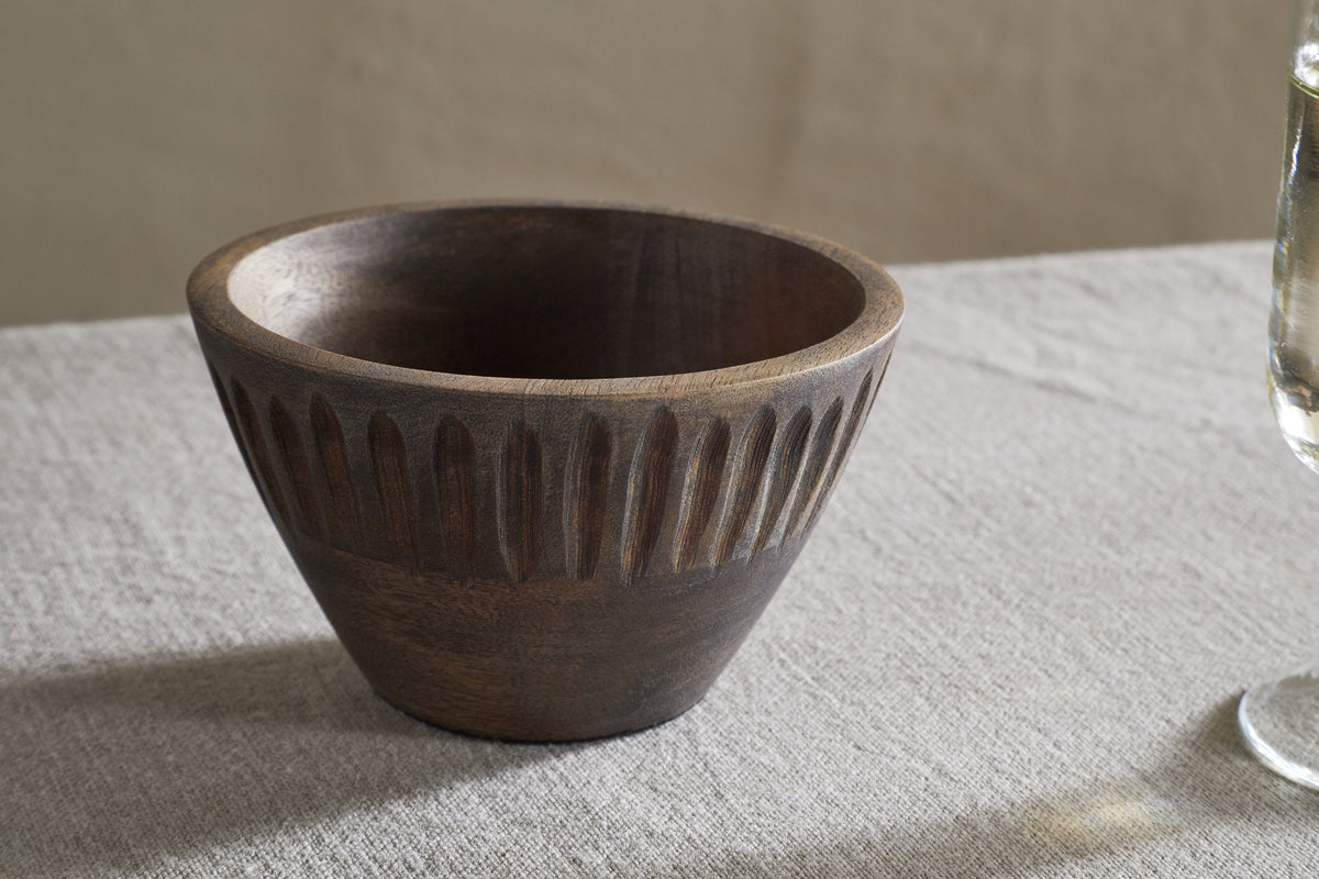 The Eisha Serving Bowl - Brown by nkuku, featuring vertical carved grooves near the rim, sits on a light beige textured tablecloth with a clear drinking glass partially visible to the right against a softly blurred neutral background.