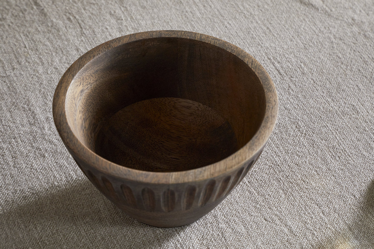 The Eisha Serving Bowl - Brown by nkuku is a round, empty decorative wood bowl with a smooth interior and vertical grooves. Featuring a rich, dark brown finish, it sits on a light beige fabric surface and makes an elegant accent piece.