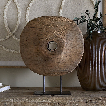 The Cala Reclaimed Wood Objet on Stand - Natural by nkuku features a rustic wooden disc with a central hole on a black metal base, displayed beside a ribbed brown vase, green branches, abstract wall art, and two stacked books.