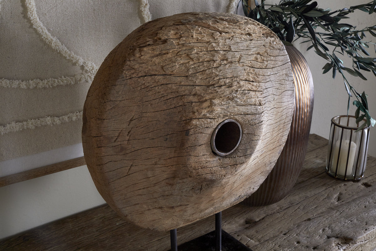 The Cala Reclaimed Wood Objet on Stand - Natural by nkuku stands upright, featuring a weathered disc with an off-center hole. Behind, a brown vase with green leaves and a glass candle holder complete the rustic heritage display.