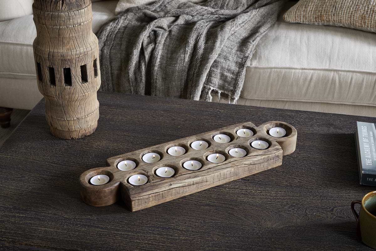 The nkuku Makala Reclaimed Wood Tealight Holder - Natural, with fourteen glowing candles, rests on a dark wooden coffee table. A tall carved decor piece, grey textured throw on a beige sofa, and The Legend of Lythmus book complete the look.