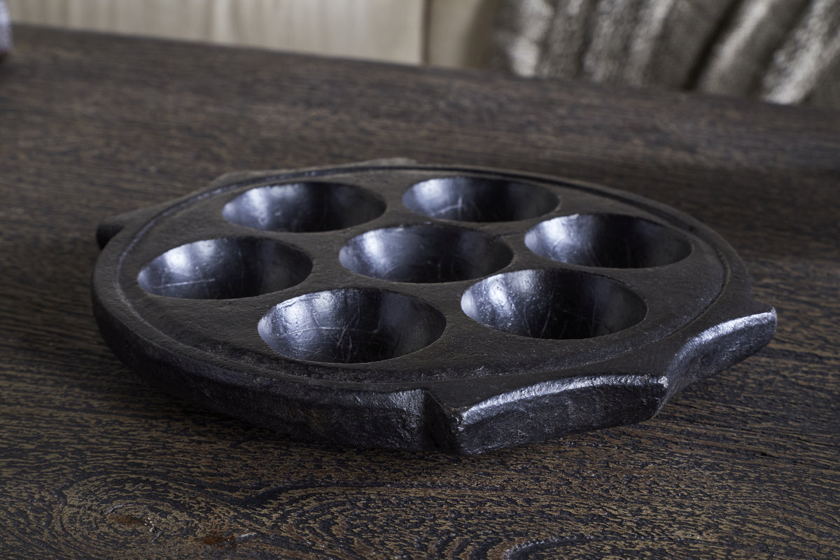The nkuku Maji Stone Multi Tealight Holder in Antique Black rests on a dark wood table, serving as a sculptural centerpiece or functional décor. In the background, a blurred sofa and textured cushion add to the stylish setting.