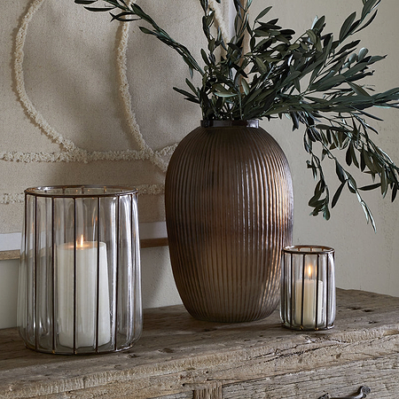A rustic wooden console displays a large ribbed brown vase with olive branches, antique brass tealight holders, and the nkuku Hava Lantern - Clear with a lit pillar candle. Textured beige wall art hangs in the background.