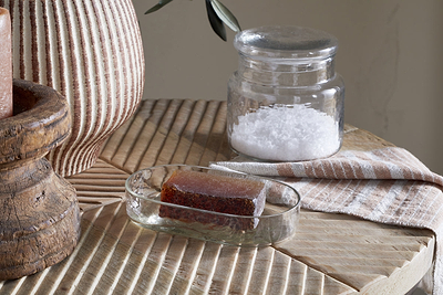 A rectangular brown soap rests in the Ilcoso Recycled Hammered Glass Soap Dish by nkuku on a wooden surface. Nearby are a jar of white bath salts, a striped cloth, and a ribbed ceramic vase in soft, neutral tones.