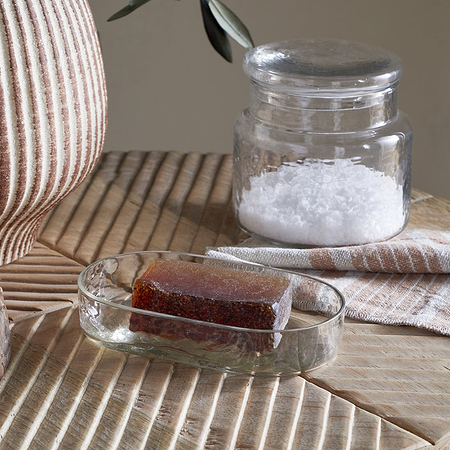 A rectangular brown soap rests in the Ilcoso Recycled Hammered Glass Soap Dish by nkuku on a wooden surface. Nearby are a jar of white bath salts, a striped cloth, and a ribbed ceramic vase in soft, neutral tones.