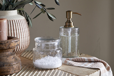 The Ilcoso Recycled Hammered Glass Storage Pot by nkuku sits on a wooden surface beside a brass-pump soap dispenser, striped towel, ribbed ceramic vase with greenery, and rustic candle holder, all under warm bathroom lighting.