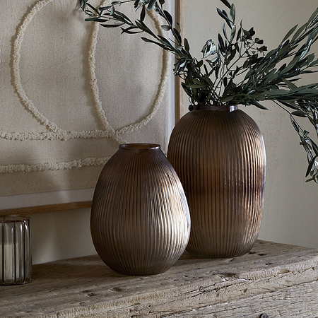 The Linsy Ribbed Glass Vase in Smoke Brown by nkuku, paired with a shorter clear vase filled with green branches, sits on a rustic wooden table alongside a candle holder, book, and textured wall art for minimalist, earthy decor.