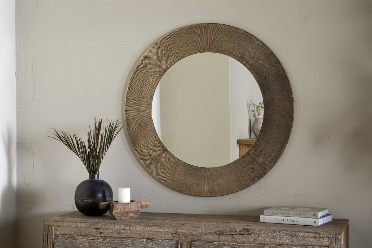 The nkuku Moto Round Mirror in Antique Brass hangs above a rustic wooden console table. On the table are a dark vase with tall green leaves, a white candle on a wooden tray, and two stacked books. The mirror reflects minimalist decor.