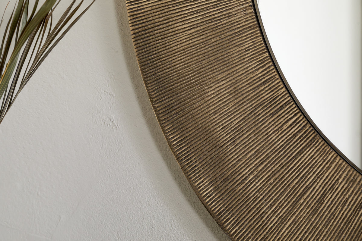 The Moto Round Mirror - Antique Brass by nkuku features a textured, grooved frame. In this close-up, it reflects a plain white wall, with thin brown plant leaves accenting the left edge for a minimalist look.