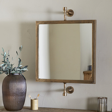 The nkuku Visala Swivel Wall Mirror in Antique Brass is mounted on a beige wall above a wooden table holding a ribbed brown vase with green branches, a gold brush cup, glass container, and metallic candle holder.