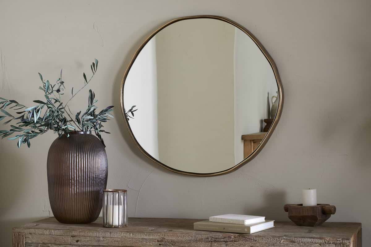 The Mchanga Organic Shape Mirror by nkuku, featuring an antique brass frame, hangs on a beige wall above a rustic wooden table with a ribbed brown vase of greenery, glass candle holder, stacked books, tray, and white candle.