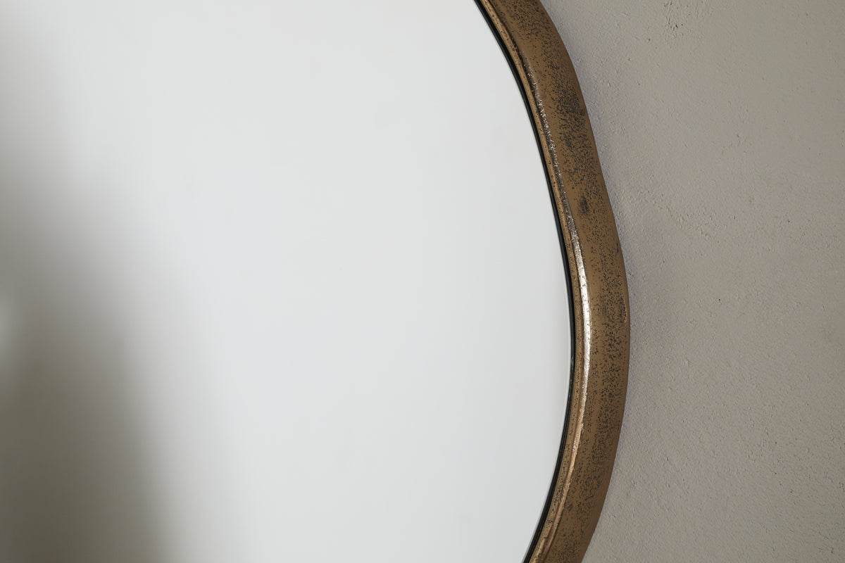 Close-up of the nkuku Mchanga Organic Shape Mirror in antique brass, mounted on a textured, light wall. The frame has a slightly weathered look, and the round mirror reflects a bright, neutral background.
