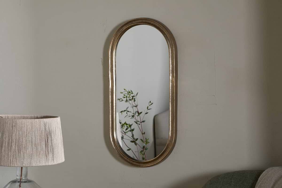 The Mbwa Oval Mirror in Antique Brass by nkuku hangs on a light gray wall. A tall lamp with a beige pleated shade stands to the left, while a light green sofa is partly visible on the right. The mirror reflects a green leafy plant.