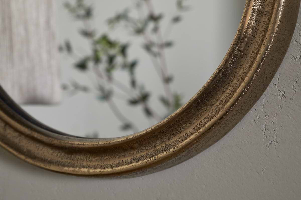 Close-up of the nkuku Mbwa Oval Mirror in antique brass, featuring an aged gold-toned frame on a light textured wall, reflecting leafy greenery and a beige curtain for a calm, relaxed ambiance.