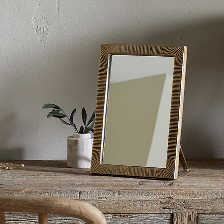 The nkuku Baja Standing Mirror - Antique Brass stands on a rustic wooden table beside a potted plant, a jewelry box with scattered jewelry, and a brown lamp with woven shade. A wooden chair and knitted blanket are in the foreground.
