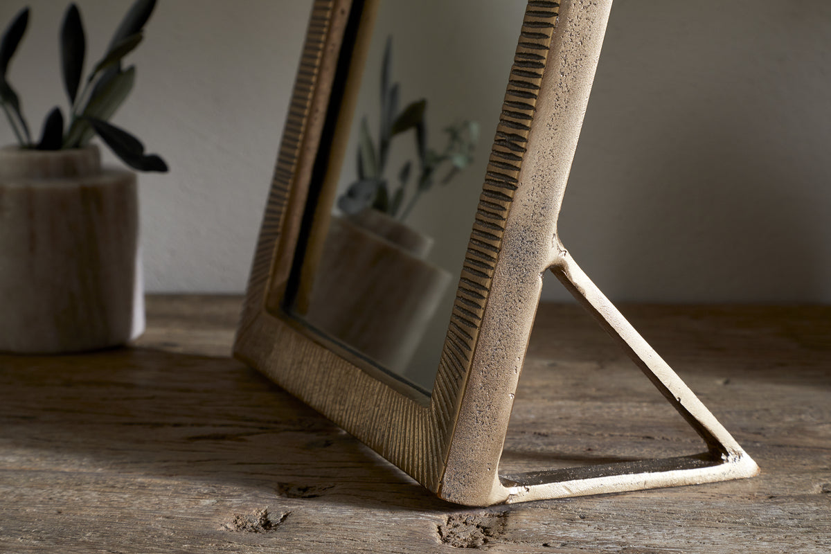 The nkuku Baja Standing Mirror in Antique Brass, featuring a textured frame and triangular stand, sits on a rustic wooden surface. Its reflection captures a blurred plant in a marble pot, softly lit by natural light.