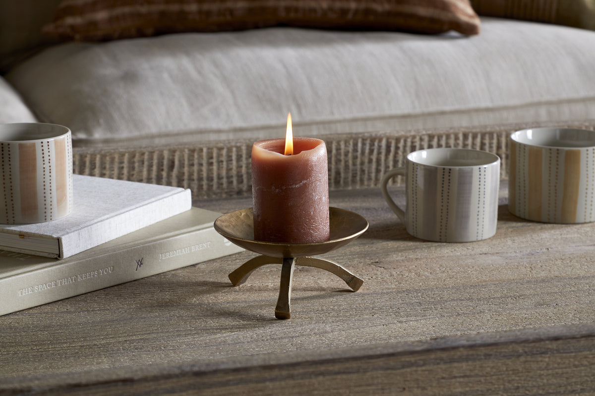 A brown pillar candle rests on the nkuku Kalik Candle Holder in Antique Brass, placed on a wooden table beside two beige-patterned mugs and a white book. In the background, a beige sofa is adorned with cushions and a woven blanket.