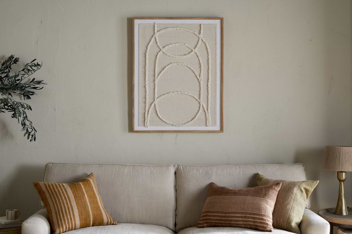 A beige sofa with two striped cushions sits against a light wall. Above hangs the Mauri Frayed Wall Art - Neutral by nkuku, its textured, curved lines framed. Side tables with a lamp and plant complete the cozy, neutral-toned decor.