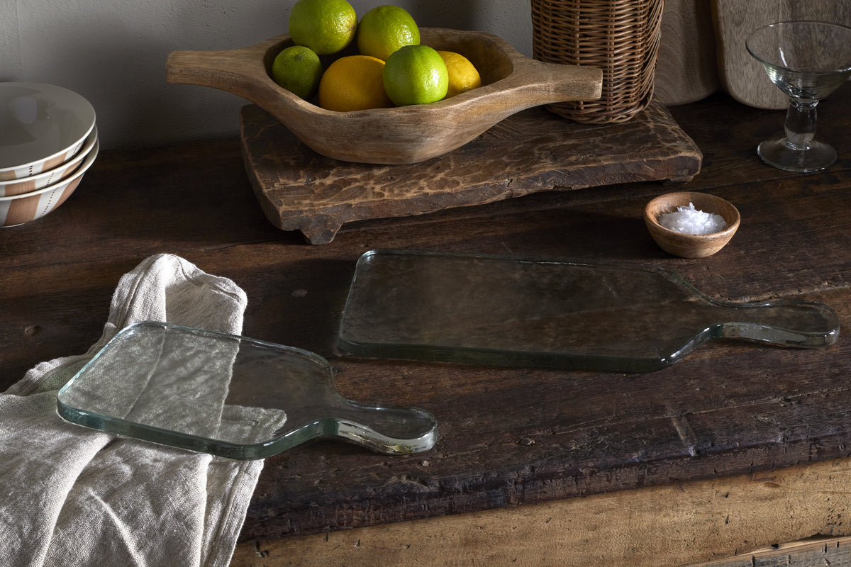 Two nkuku Komal Serving Platters in clear glass with handles rest on a rustic wooden table. One sits atop a beige cloth. Nearby are bowls, a wooden fruit bowl with lemons and limes, a wicker basket, and serveware.