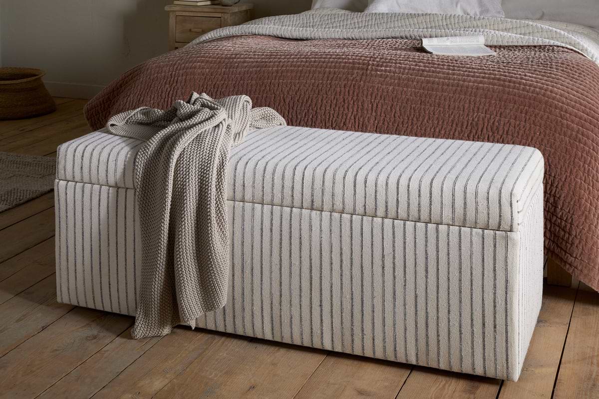 The nkuku Sanja Stripe Cotton & Linen Upholstered Storage Bench in Grey sits at the foot of a bed with a textured reddish-brown quilt and white sheets, adding practical bedroom storage to the cozy, minimalist space with neutral decor and wooden floors.