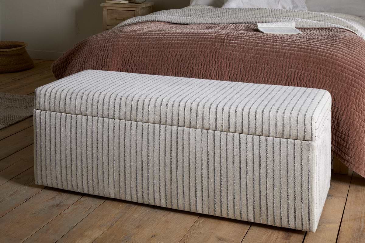 The nkuku Sanja Stripe Cotton & Linen Upholstered Storage Bench - Grey, featuring vertical beige and grey stripes, adds stylish storage at the foot of the bed with mauve-brown quilt and white bedding in a cozy bedroom.