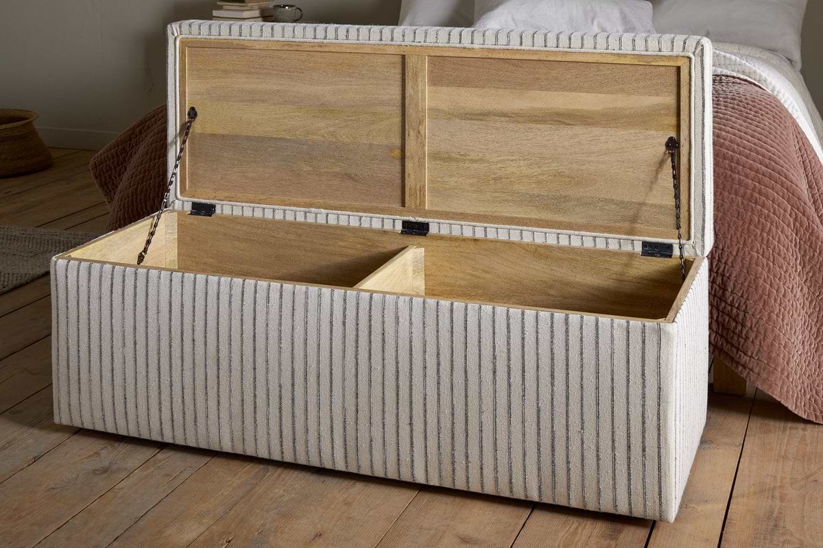 The nkuku Sanja Stripe Cotton & Linen Upholstered Storage Bench in grey features a hinged lid, two wooden compartments, and light fabric with thin gray stripes, offering stylish bedroom storage beside a bed.