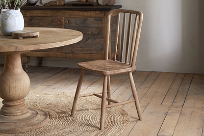 The nkuku Om Mango Wood Dining Chair - Natural, featuring vertical slats, sits on a round woven rug beside a matching table, with a wooden floor and sideboard setting the scene for a cozy farmhouse-style interior.