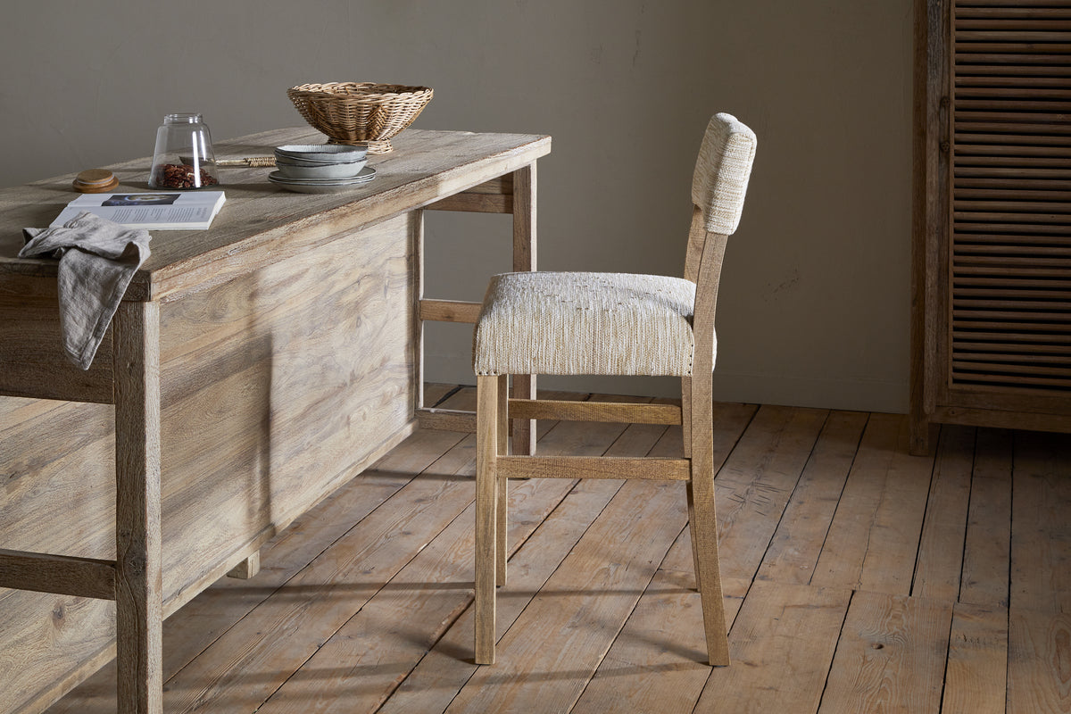 A rustic mango wood table with dishes, a glass jar, and a basket sits on a wooden floor. The Zoha Cotton & Jute Upholstered Counter Chair by nkuku is angled toward the table in a softly lit, minimalist room.
