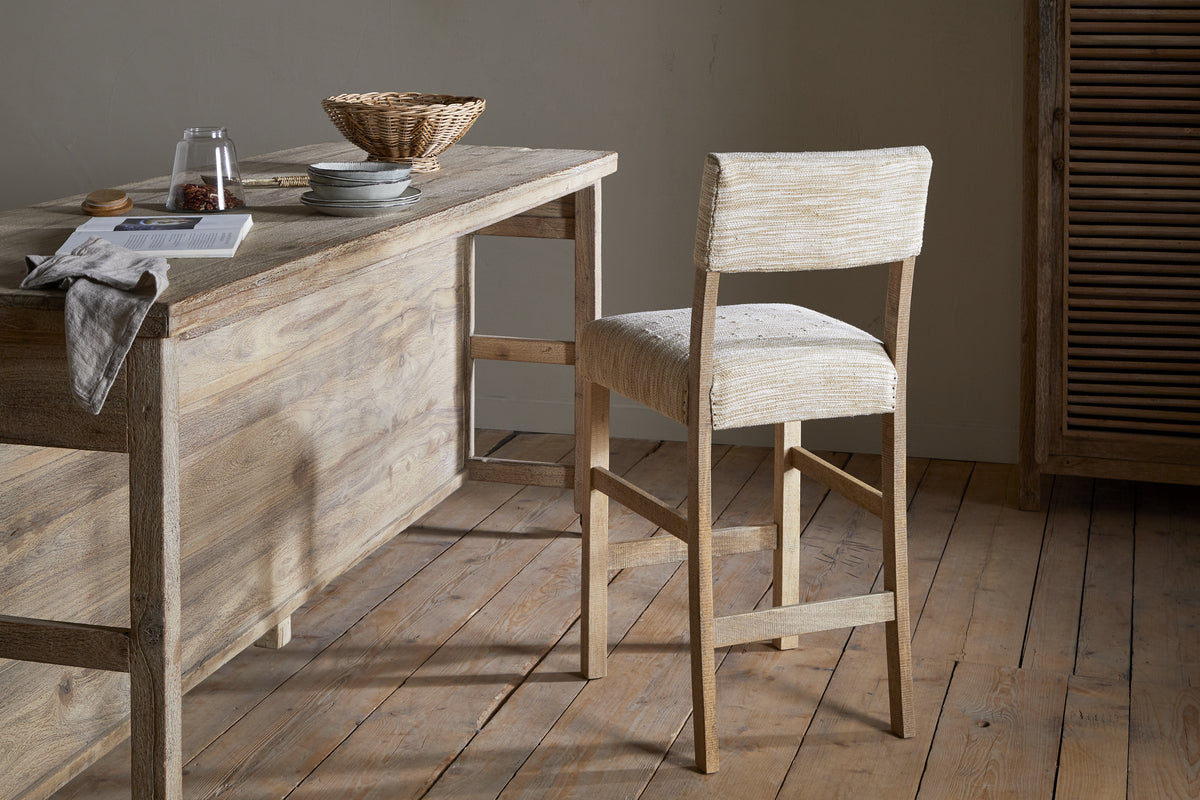 The Zoha Cotton & Jute Upholstered Counter Chair by nkuku stands beside a rustic mango wood bar-height table on a light floor. Sunlight warms the cozy room, where snacks and drinks sit atop the table in woven and ceramic accents.