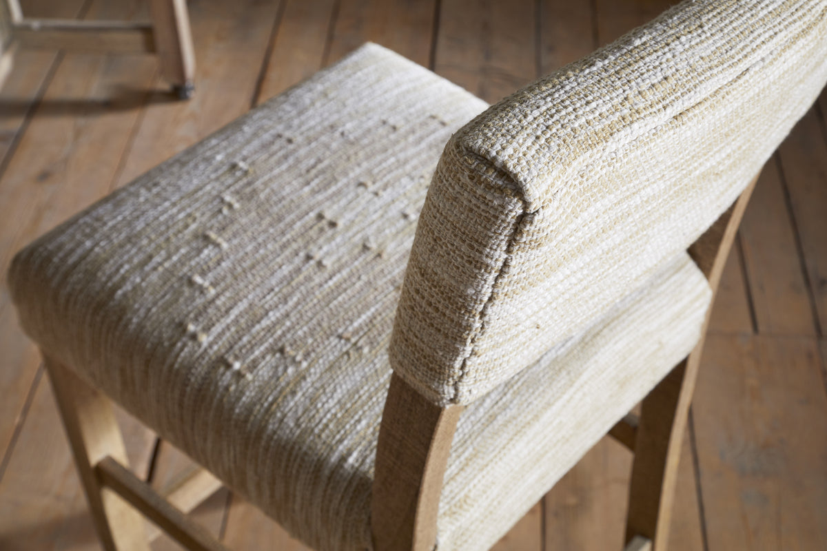 A close-up of the nkuku Zoha Cotton & Jute Upholstered Counter Chair in Natural shows its textured cream seat and backrest, with sunlight highlighting the fabric and stitching. Another Zoha chair is partially visible in the background.