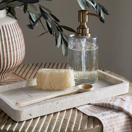 The nkuku Ganda Travertine Vanity Tray in Cream holds a natural loofah, wooden spoon, and clear soap dispenser with gold pump. It sits on a ribbed wooden table next to a textured vase with greenery and a striped cloth draped nearby.