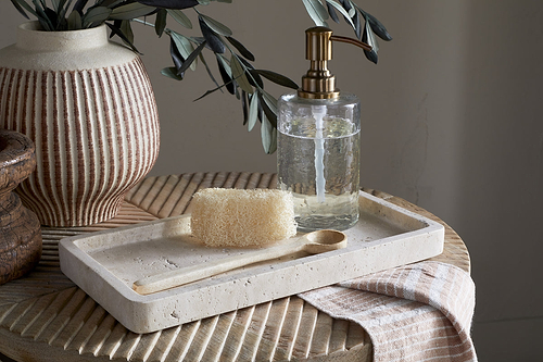 The nkuku Ganda Travertine Vanity Tray in Cream holds a natural loofah, wooden spoon, and clear soap dispenser with gold pump. It sits on a ribbed wooden table next to a textured vase with greenery and a striped cloth draped nearby.