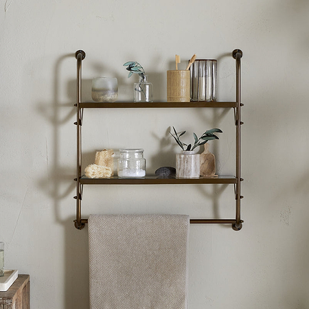 The Karura Wall Shelves with Towel Rail in Antique Brass by nkuku hold jars, a toothbrush holder, cups, a loofah, stones, and small plant cuttings. A beige towel hangs below; to the left sit a mirror and soap dispenser on a wooden counter.
