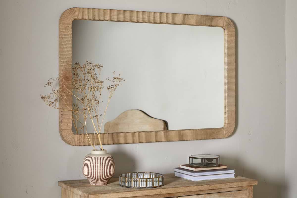 The nkuku Vivan Rectangular Grooved Wood Mirror in natural finish hangs above a wooden dresser topped with a striped vase of dried flowers, round tray, glass box, and books, set against a plain light-colored wall.