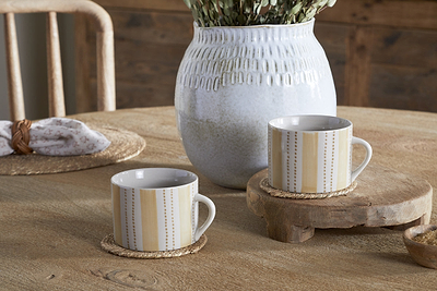 Two Patthar Mugs in Cream & Sand by nkuku rest on round coasters atop a wooden table, with a white textured vase of dried flowers behind. A woven placemat and napkin with wooden ring complete the rustic, cozy setting. Set includes 2 large mugs.