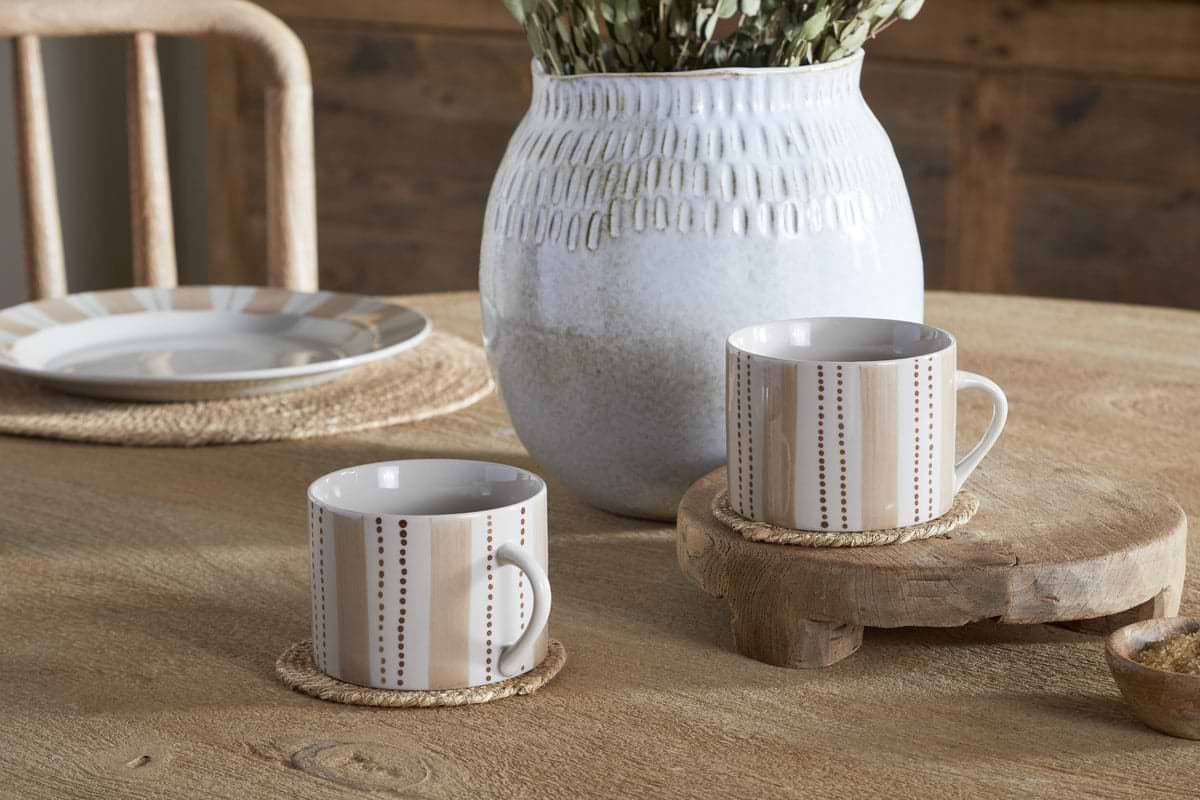 Two large nkuku Patthar Mugs in Cream & Clay rest on round woven coasters atop a wooden table—one elevated on a rustic stand. A white patterned vase with greenery and a striped-rim Patthar plate complete the setting.