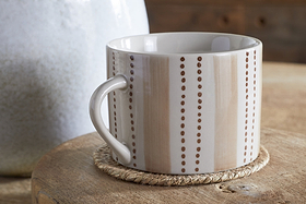 The Patthar Mug - Cream & Clay - Large (Set of 2) by nkuku, featuring beige and white stripes and dotted lines, rests on a woven coaster atop wood. A large white vase in the background adds to the cozy, rustic vibe.