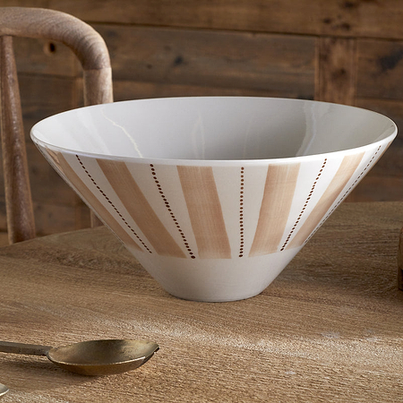 The nkuku Patthar Serving Bowl - Cream & Clay - Medium, hand-painted with beige stripes and dotted lines, rests on a wooden table. A gold spoon with a wrapped handle and a textured plant pot add rustic dinnerware accents.