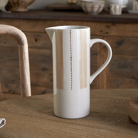 The nkuku Patthar Jug - Cream & Clay - Large, a tall hand-painted ceramic piece with delicate vertical dotted lines, sits on a wooden table set with a napkin, woven placemat, and large vase. A rustic chair and sideboard complete the cozy scene.