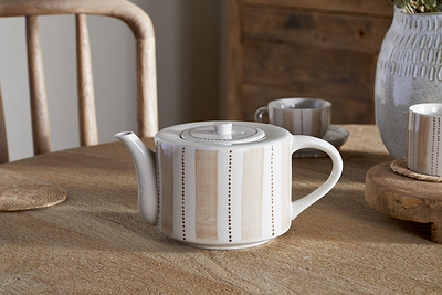The nkuku Patthar Teapot – Cream & Clay – Large, featuring hand-painted stripes and dots in beige, sits on a wooden table with its matching cup, woven coaster, gray vase with greenery, and wooden chairs in the background.