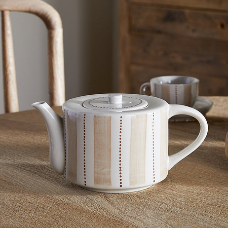 The nkuku Patthar Teapot – Cream & Clay – Large, featuring hand-painted stripes and dots in beige, sits on a wooden table with its matching cup, woven coaster, gray vase with greenery, and wooden chairs in the background.
